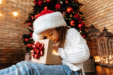 Christmas. Childhood. Presents. Home. Happy little girl is opening a gift while sitting near the Xmas tree
