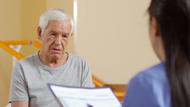 Medium shot of senior man with grey hair answering questions of female physical therapy specialist making notes on clipboard