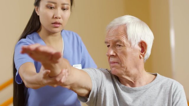 Medium shot of young female physical therapist moving arm of elderly man and asking if he is feeling any pain