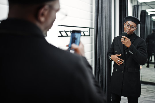 Stylish casual african american man at black beret and overcoat at fitting room clothes store, making photo at mirror on his phone camera.