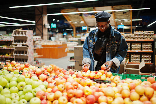 Stylish casual african american man at jeans jacket and black beret checking apple fruits in organic section of supermarket.