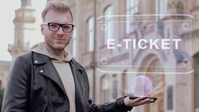 Smart young man with glasses shows a conceptual hologram E-ticket. Student in casual clothes with future technology mobile screen on university background