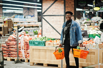 Stylish casual african american man at jeans jacket and black beret holding two baskets, walking and shopping at supermarket.