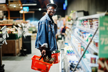 Stylish casual african american man at jeans jacket and black beret holding two baskets, standing near fridge and shopping at supermarket.
