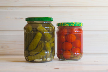 Salted red tomatoes and green gherkins in jars on a light wooden background.