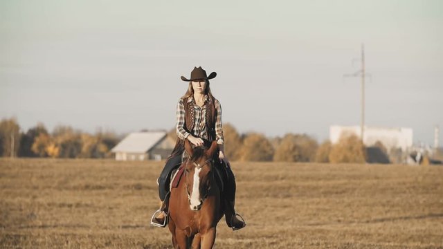 Beautiful woman riding horse in background sunrise in field. Young cowgirl at brown horse in slow motion outdoors. Medium shot
