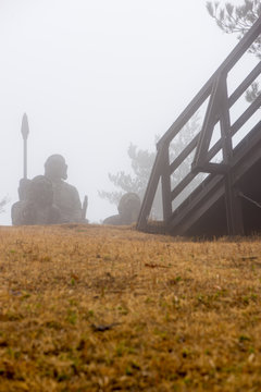 Statues of ancient indigenous peoples atop a hill next to a bridge on a foggy day. Vertical orientation. Kunimigaoka peak, Mount Aso. Takachiho, Miyazaki, Japan. Travel and nature.