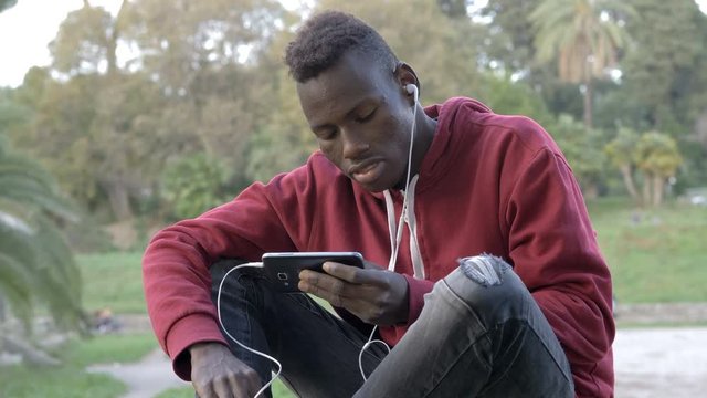 Young american african man sitting on lawn listening music with smartphone