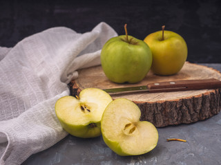 Two green golden apples and two halves on a wooden platter and gray background, cotton white napkin, kitchen knife