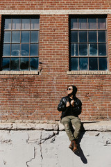 Young Attractive Modern Fashion Caucasian Male Guy Walking, Sitting, Smiling, and Laughing Outside Urban Old Abandoned Brick Warehouse Building in Urban City in Winter Season on Bright Sunny Day