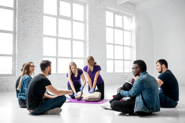 Fototapeta premium Group of young people sitting in a circle during the first aid courses with two instructors training on medical dummies in the white spacious room