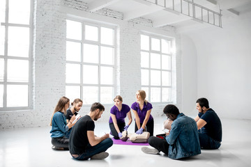 Fototapeta premium Group of young people sitting in a circle during the first aid courses with two instructors training on medical dummies in the white spacious room