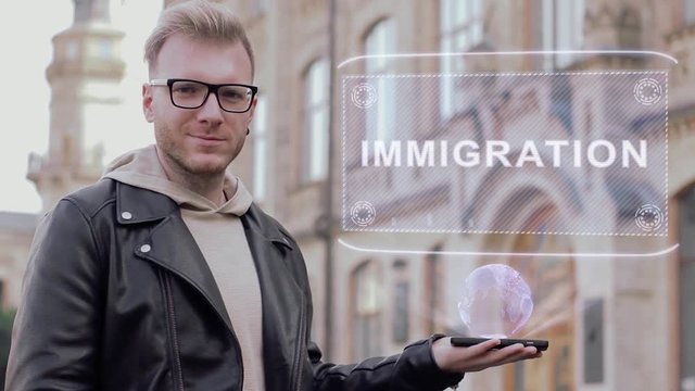 Smart young man with glasses shows a conceptual hologram Immigration. Student in casual clothes with future technology mobile screen on university background