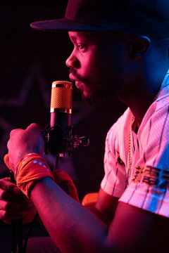 Studio portrait of young man with beard and mustaches in baseball cap and t-shirt with microphone. Pink neon light