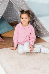 beautiful happy child sitting on carpet near teepee and smiling at camera