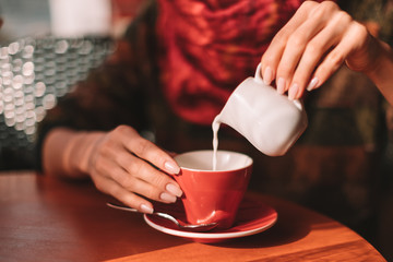 Close up of Woman’s hands pouring milk in red coffee cup - woman sitting in cafe with soft drink