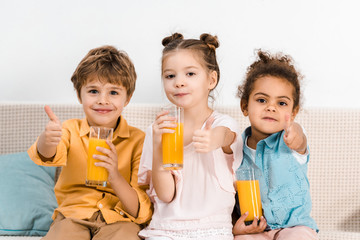 cute multiethnic kids holding glasses of juice and showing thumbs up