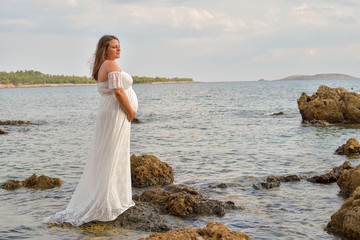 Fototapeta na wymiar pregnant woman in white dress sitting on rocks on the beach
