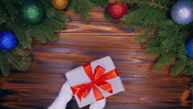 Wooden table with Christmas decorations. Cat paws giving gift wrapped in silver shining paper and red bow. Top plan view. Animal, pets, new year concept. Slow motion.