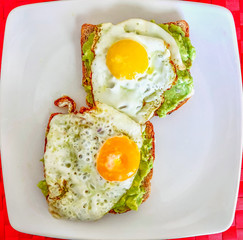 two fried eggs on avacado guacamole and brown toast on a white square shape plate, red place mat from above