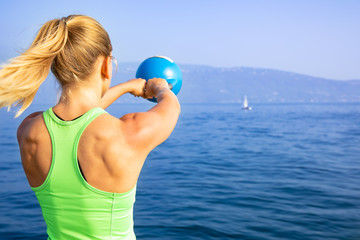 young sexy woman are doing some cross workout with a kettlebell to get fit at a summer day in the sun at the water