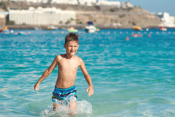 The portrait of an active European boy in a stripes swimming shorts.  He is running from the sea, smiling and enjoying his summer vacations.
