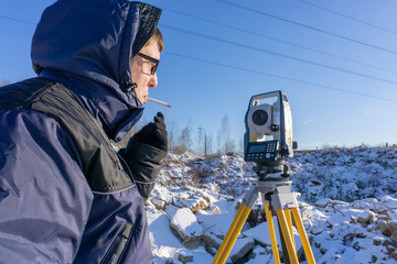 Surveyor with a cigarette in his mouth conducts a topographical survey for the inventory at a construction site in the winter