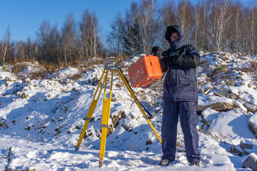 Surveyor carries a geodetic instrument in an orange case and a yellow tripod at a construction site in winter
