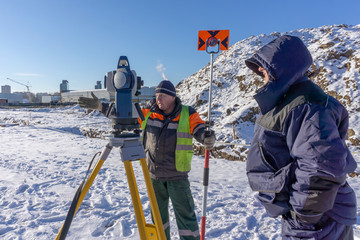 A surveyor with an assistant makes a topographical survey for the cadastre at a construction site in winter