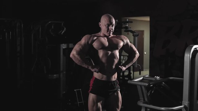 Bodybuilder posing and demonstrating chest muscles and arms in dark gym. Young strong male bodybuilder flexing muscles in slow motion