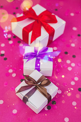 Three gift boxes with colorful ribbons on pink background with confetti.