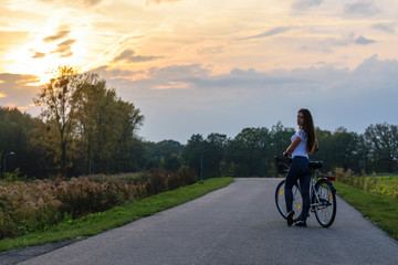 Obraz premium Young brunette girl with a bicycle in a park alley lit by sunset light.