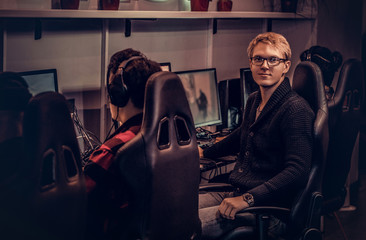A young smart gamer wearing a sweater and glasses sitting on a gamer chair and looking at a camera in a gaming club or internet cafe.
