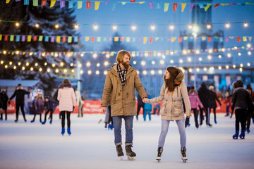 Young couple in love Caucasian man with blond hair with long hair and beard and beautiful woman have fun, active date skating on ice scene in town square in winter on Christmas Eve
