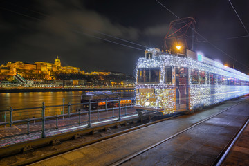 Budapest, Hungary - Festively decorated light tram (fenyvillamos) on the move with Buda Castle Royal Palace at Vigado square by night. Christmas season in Budapest
