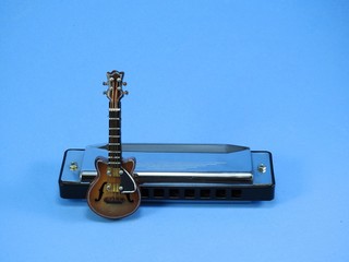 A miniature brown electric guitar leaning on a silver 10-hole diatonic harmonica. Blue background.