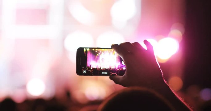 Berlin, Germany - 14 August 2018: Back shot of young man holding smartphone in hand. Fan making video at concert. Outdoors. Colored lights. Outdoors.