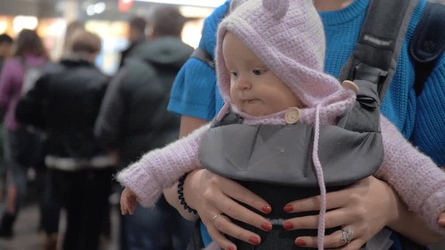 Medium panoramic handheld shot of a cute baby in a baby carrier while waiting with her mother to travel in the airport terminal