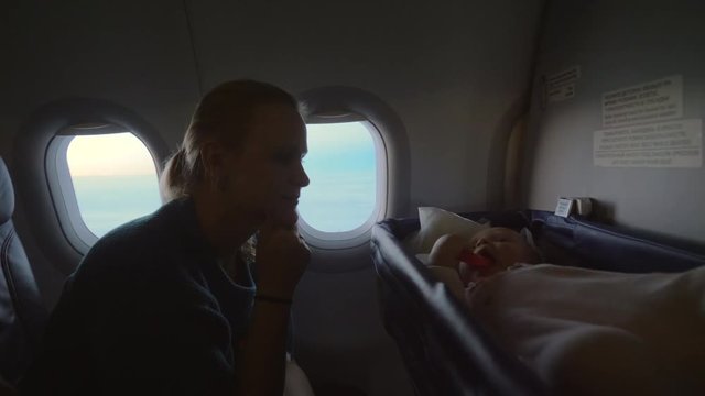Medium shot of a mother traveling with her baby on a airplane while she plays in a bassinet