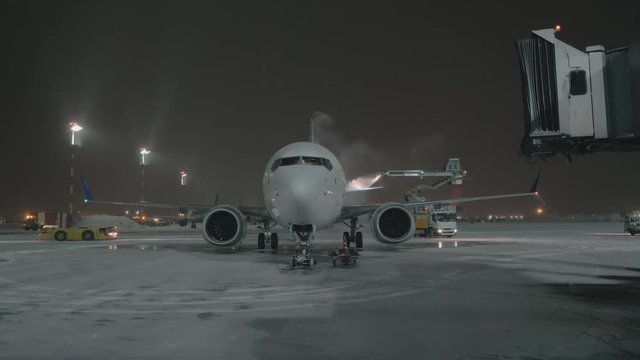 A white glossy airplane is standing on a courtyard. It is being washed by a heavy water jet from a technical car with an elevator. There is some snow on the ground and the sky is dark. Many different