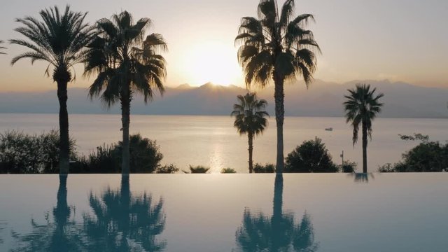A steadicam shot of an open pool glossy water boundary. There are tall dark palm trees behind it and a picturesque sea scenery. Misty mountain hills are filling the background and warm rays of bright