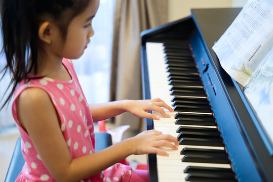 Young cute Asian kid girl playing electric piano at home. She is practicing piano after she had piano class at school. Seen from side of the girl. Happy piano playing concept.