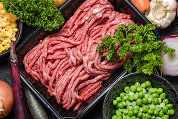 Raw minced meat in container with ingredients for shepherds pie with green peas, yellow corn, carrot, onion and seasonings, on black background, top view