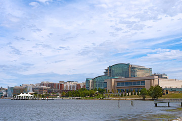 National Harbor waterfront panorama in Oxon Hill, Maryland, USA. Sun shines through cumulus clouds on National Harbor pier and modern buildings along coastline of Potomac River.