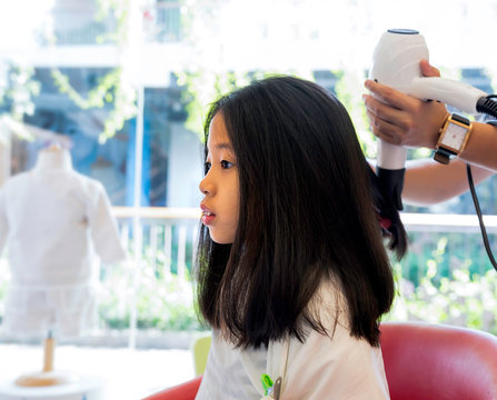 Portrait Of Asian Kid With Healthy Long Black Hair Getting Haircut. Hand Of Stylist Drying Woman Hair In Beauty Salon With Blower. Hairdresser Making Hairstyle To Pre Teen Girl In Barber Shop.