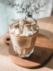 One glass of hot coffee with marshmallow on top serve on white tray on wood table next to bright window in morning.