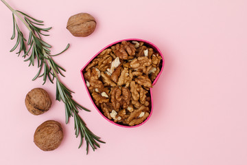Peeled walnuts in gift box in shape of heart with unpeeled ones and rosemary on a pink background.