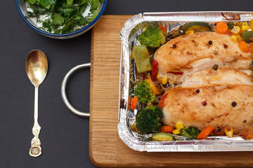Baked chicken breasts or fillet in metal container with vegetables on cutting board.
