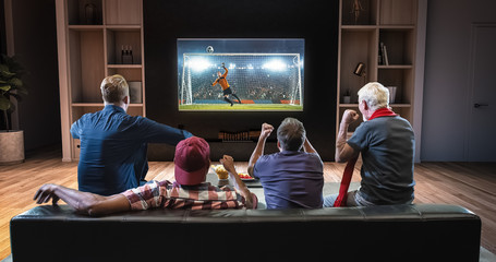 Group of fans are watching a soccer moment on the TV and celebrating a goal, sitting on the couch in the living room.