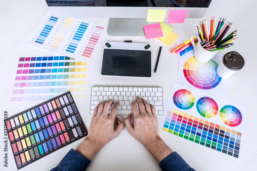 Poster Image of male creative graphic designer working on color selection and drawing on graphics tablet at workplace with work tools and accessories, top view workspace
